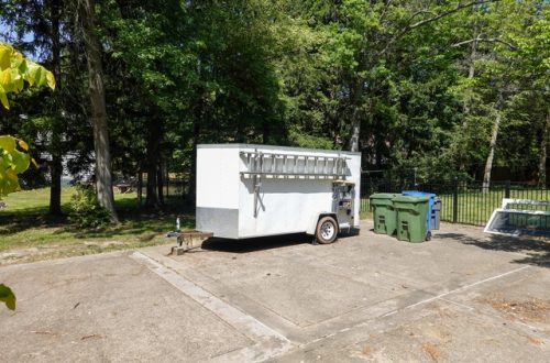 A white storage trailer parked in the driveway next to two green garbage cans. The trailer has a silver ladder on the side.
