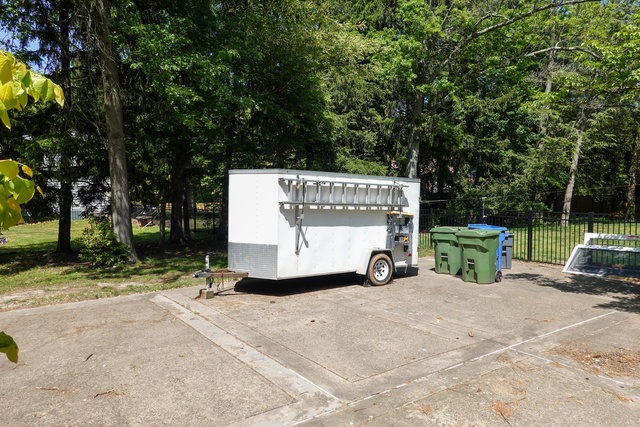 A white storage trailer parked in the driveway next to two green garbage cans. The trailer has a silver ladder on the side.
