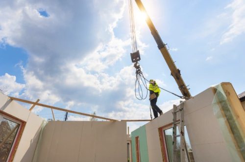 A construction worker walking along the unfinished parts of a house while guiding the hook of a crane to a certain spot.