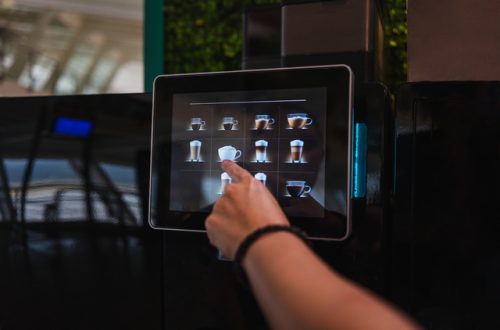 A close-up view shows a person pressing the choice of coffee on the screen of a coffee machine. The area is dimly lit.