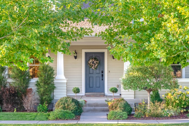 The exterior of a home features a blue front door, a small front porch, green trees, and beige siding.