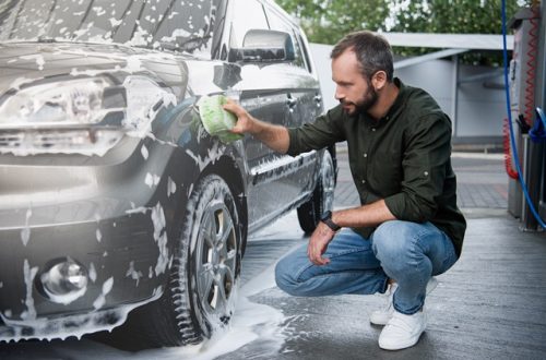 A man squatting next to his car with a sponge in hand as he washes the vehicle. The car is covered in soap.