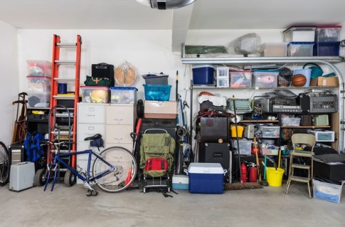 A room with many items stacked up against a white wall. There are boxes on shelves and a ladder off to the side.