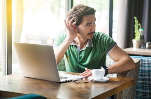A man sitting at a wooden table holds a hand to his ear with a puzzled look on his face. A mug and a laptop are on the table.