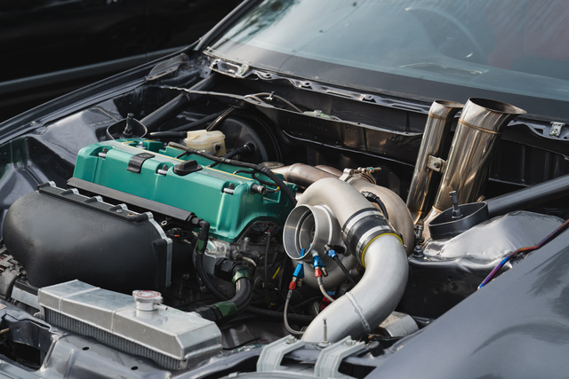 A close-up of a vehicle with its hood up and engine bay exposed. Upgrades and mods are visible, including a green valve cover.