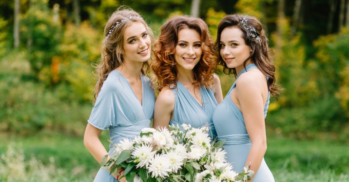 Three women holding large bouquets with white flowers are wearing light-blue bridesmaid dresses and posing.
