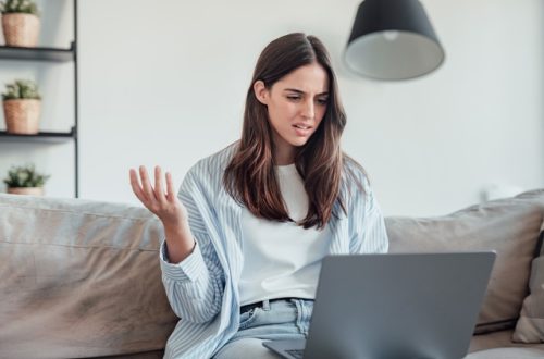 A young woman sits on a couch looking at an open laptop. She has an upset look on her face with a raised arm.