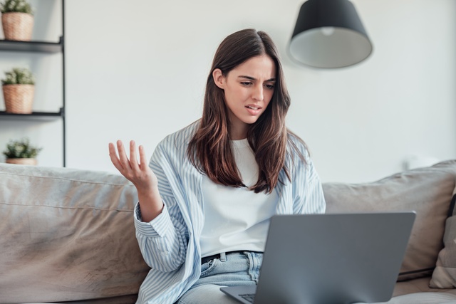 A young woman sits on a couch looking at an open laptop. She has an upset look on her face with a raised arm.
