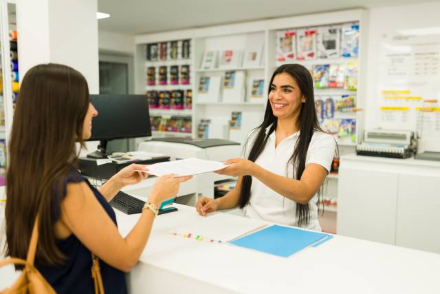 A woman wearing a white Polo shirt is smiling and handing a female customer a stack of papers from the print shop.