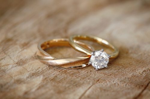 A gold wedding band and a solitaire diamond engagement ring resting together on a textured wooden table.