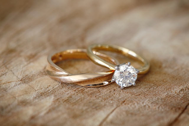 A gold wedding band and a solitaire diamond engagement ring resting together on a textured wooden table.