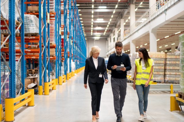 Three workers walk through a large warehouse aisle, reviewing a tablet beside tall storage racks and stacked inventory.