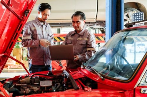 Two mechanics standing over the open hood of a car in an auto repair shop. One mechanic holds a laptop.