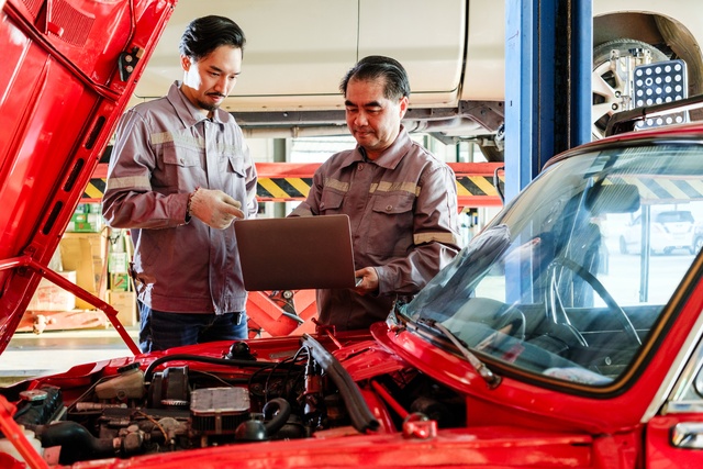 Two mechanics standing over the open hood of a car in an auto repair shop. One mechanic holds a laptop.