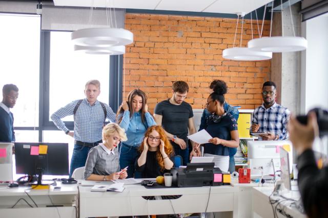 An office that is crowded with many different coworkers. Some stand around desks while two women sit at a desk.