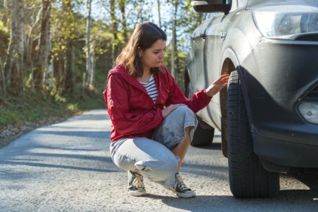 A woman in a red jacket and jeans crouches next to a parked vehicle to inspect the wheel and tire on an empty road.