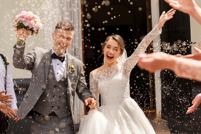 A bride and groom walk forward smiling as guests toss pieces into the air, while she holds a bouquet and raises one arm.