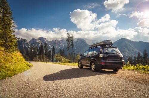 Car with rooftop carrier stopped on scenic mountain road, overlooking forest and rugged mountains under blue sky.