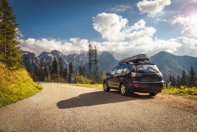 Car with rooftop carrier stopped on scenic mountain road, overlooking forest and rugged mountains under blue sky.