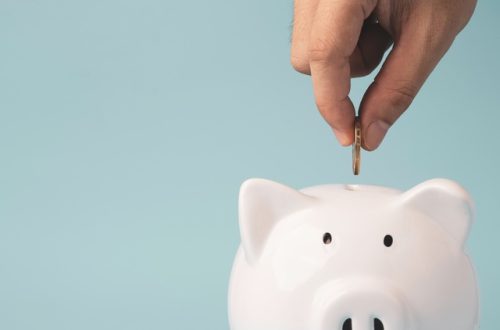 A close-up view shows a person's hand placing a coin into the slit on the top of a white piggy bank.
