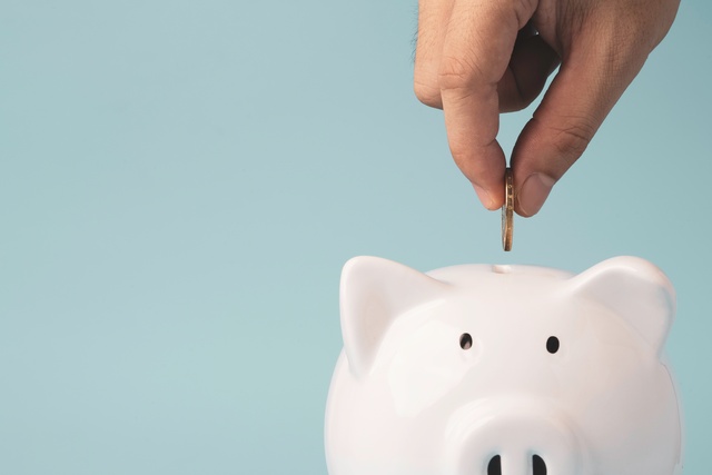A close-up view shows a person's hand placing a coin into the slit on the top of a white piggy bank.