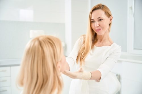 A woman beauty technician stands in front of a client sitting down in a chair, inspecting her facial features.