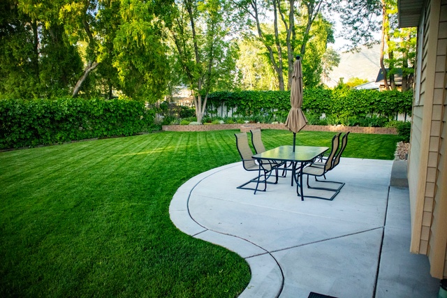 A medium-sized concrete patio features a rectangular glass outdoor table with chairs and an umbrella in a backyard.