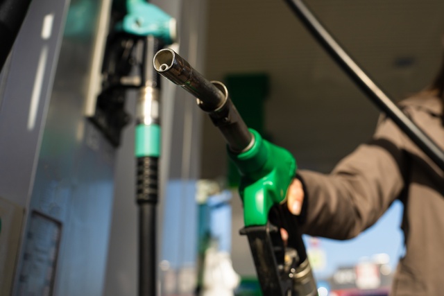 A close-up of a person in a brown sweater holding a green gasoline nozzle at a fuel station pump during golden hour.