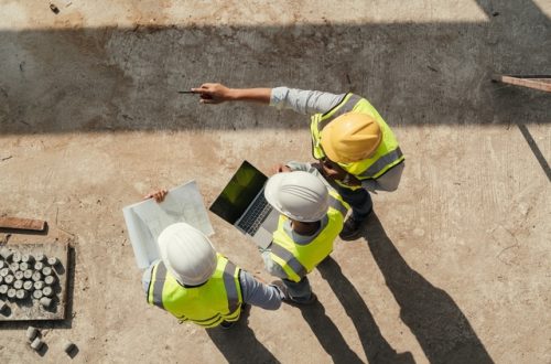 A top view of three workers standing at a construction site, looking at blueprints and a tablet with materials nearby.