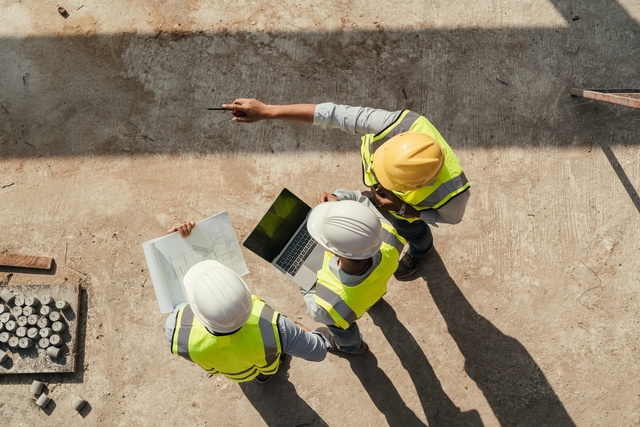 A top view of three workers standing at a construction site, looking at blueprints and a tablet with materials nearby.
