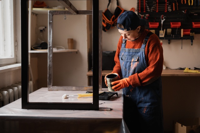 Person paints a black metal frame in a workshop, wearing overalls, gloves, and a cap near shelves and tools.