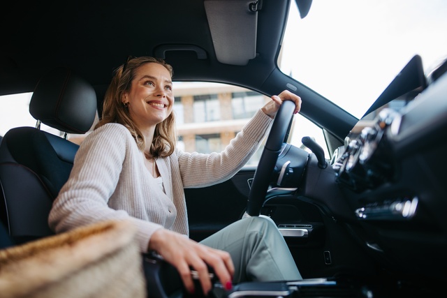 A happy woman in a sweater driving her car. She has one hand on the wheel and another on the gear shift.