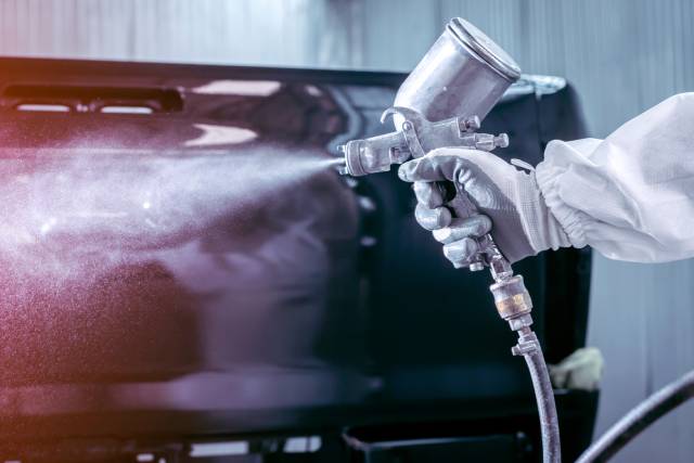 A close-up view shows a person wearing a white protective suit using a sprayer to paint the back of a truck.