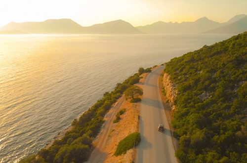 Car travels along a hillside road above a glowing sea, with dense green shrubs and hazy mountains at sunset.