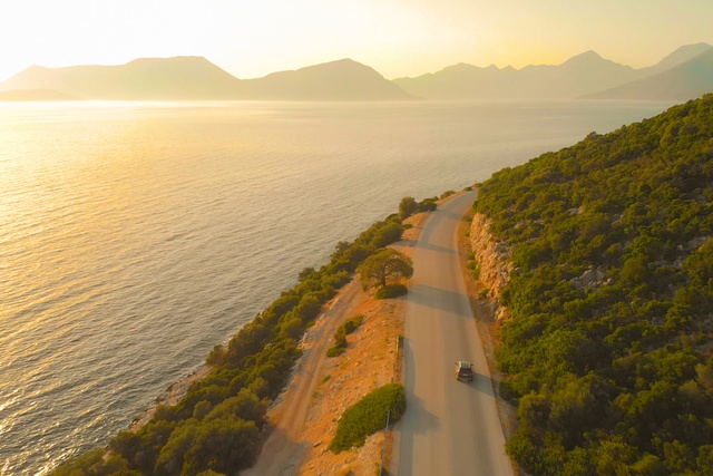 Car travels along a hillside road above a glowing sea, with dense green shrubs and hazy mountains at sunset.