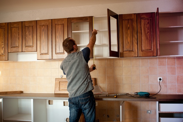 A person measuring wide wooden cabinets in a residential kitchen with a small yellow measuring tape.