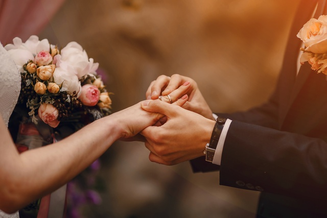 A person placing a ring on another person's finger beside a bouquet of pale flowers and a dark suit sleeve nearby.