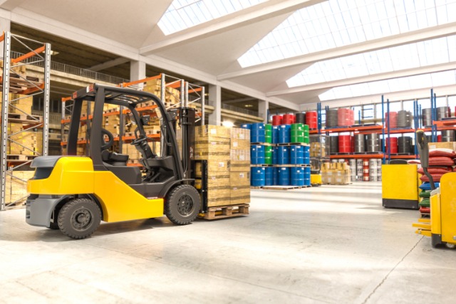 A yellow forklift lifting a pallet of wrapped boxes in a warehouse with stocked racks, blue drums, and skylights.
