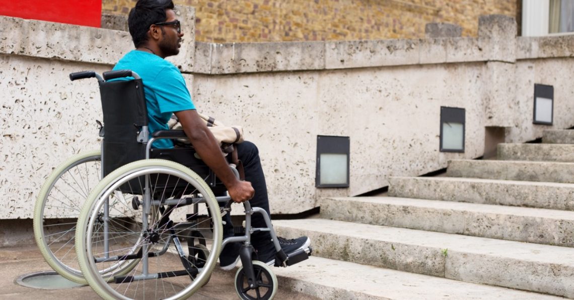 A man in a wheelchair facing concrete stairs beside a low wall, brick building, and square wall lights.