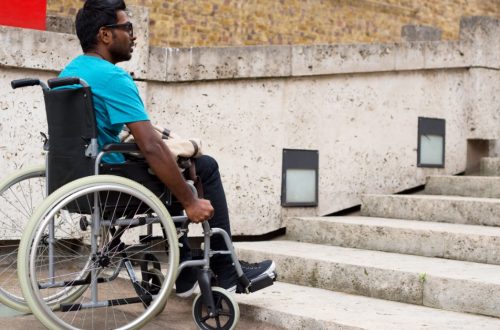 A man in a wheelchair facing concrete stairs beside a low wall, brick building, and square wall lights.