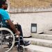 A man in a wheelchair facing concrete stairs beside a low wall, brick building, and square wall lights.