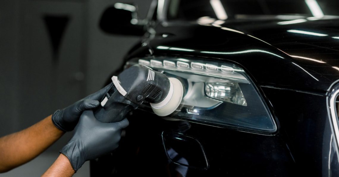 Gloved hands using an orbital polisher on a black car headlight inside a dim auto service area with bright reflections.