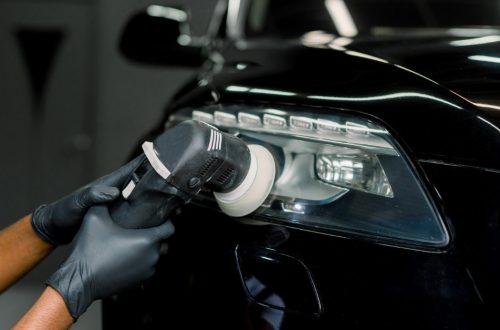 Gloved hands using an orbital polisher on a black car headlight inside a dim auto service area with bright reflections.