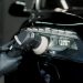Gloved hands using an orbital polisher on a black car headlight inside a dim auto service area with bright reflections.