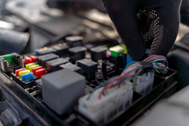 A person wearing a black glove gently grabs an electrical connector next to wires on a car fuse relay block.