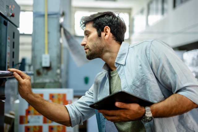 A man wearing a light-blue button-down shirt holds a table in one hand while inspecting an industrial printer with the other.