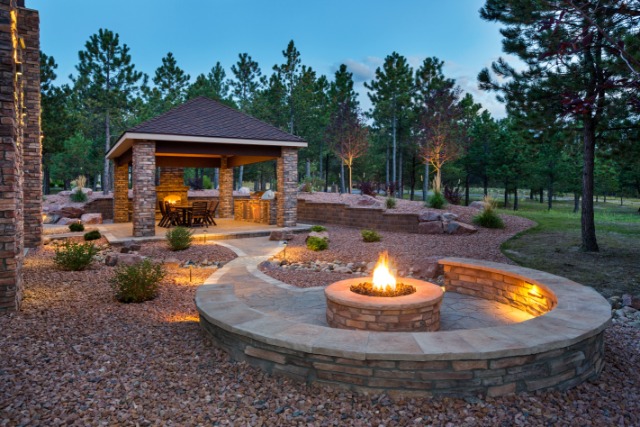 A stone patio in a large backyard. The space has a round fire pit, covered dining area, and pathway lights.