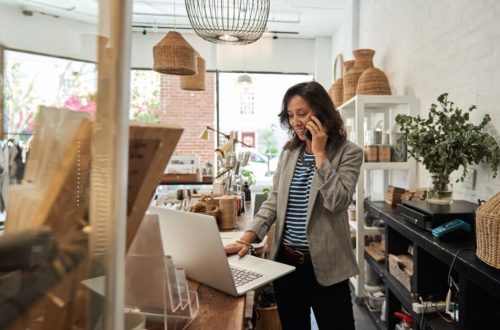 A woman smiling and talking on a phone while using a laptop at the counter of a bright home decor retail shop.