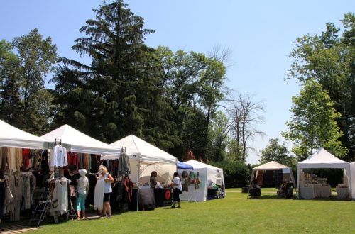 White vendor tents displaying clothing and goods as shoppers browse an outdoor market on a grassy lawn.