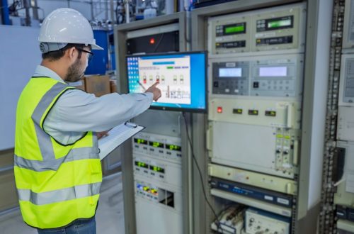 A worker wearing a yellow vest and a helmet, holding a clipboard while touching a digital screen on a control panel.